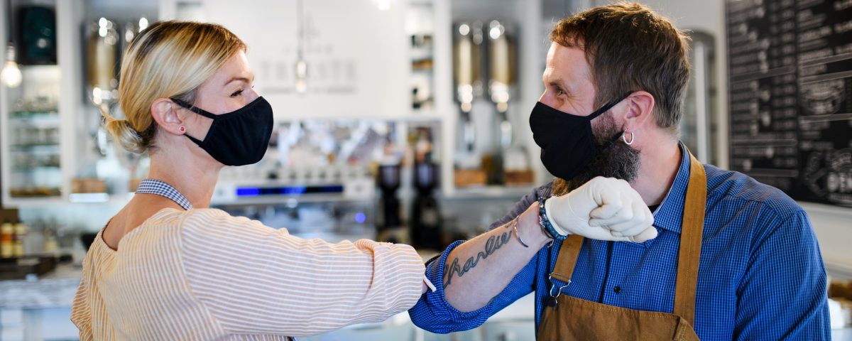 Portrait of coffee shop owners with face masks elbow bumping, open after lockdown quarantine, coronavirus concept.