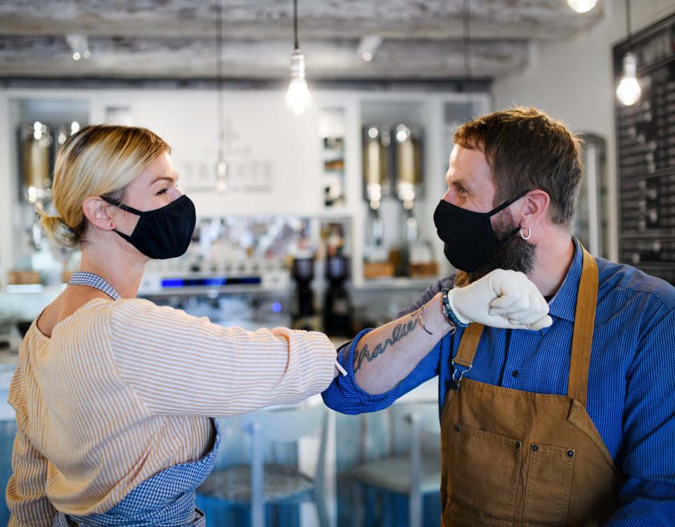 Portrait of coffee shop owners with face masks elbow bumping, open after lockdown quarantine, coronavirus concept.