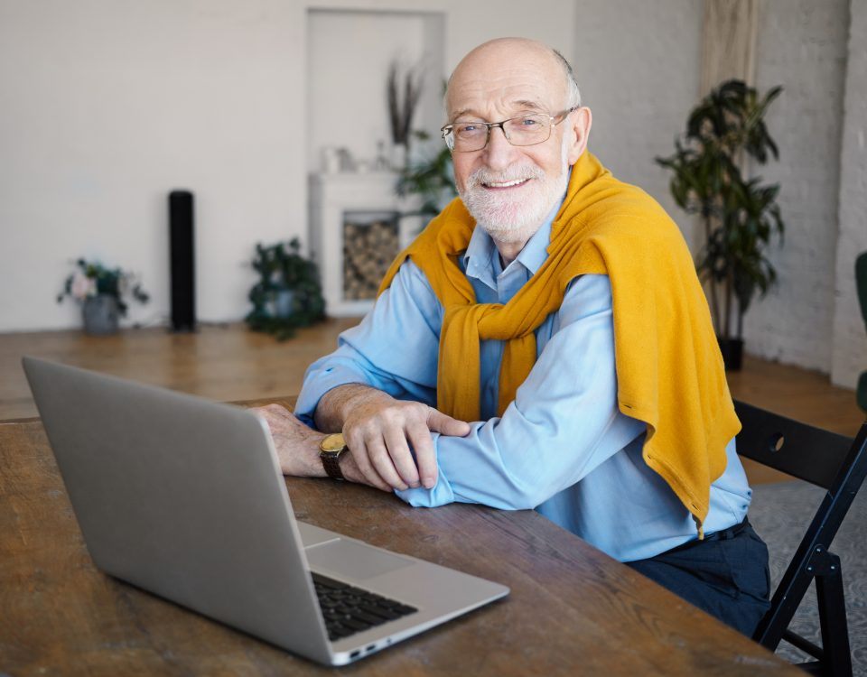 Indoor shot of handsome positive unshaven sixty year old man writer wearing eyeglasses and stylish clothes working distantly sitting at desk in front of open laptop computer, smiling broadly at camera